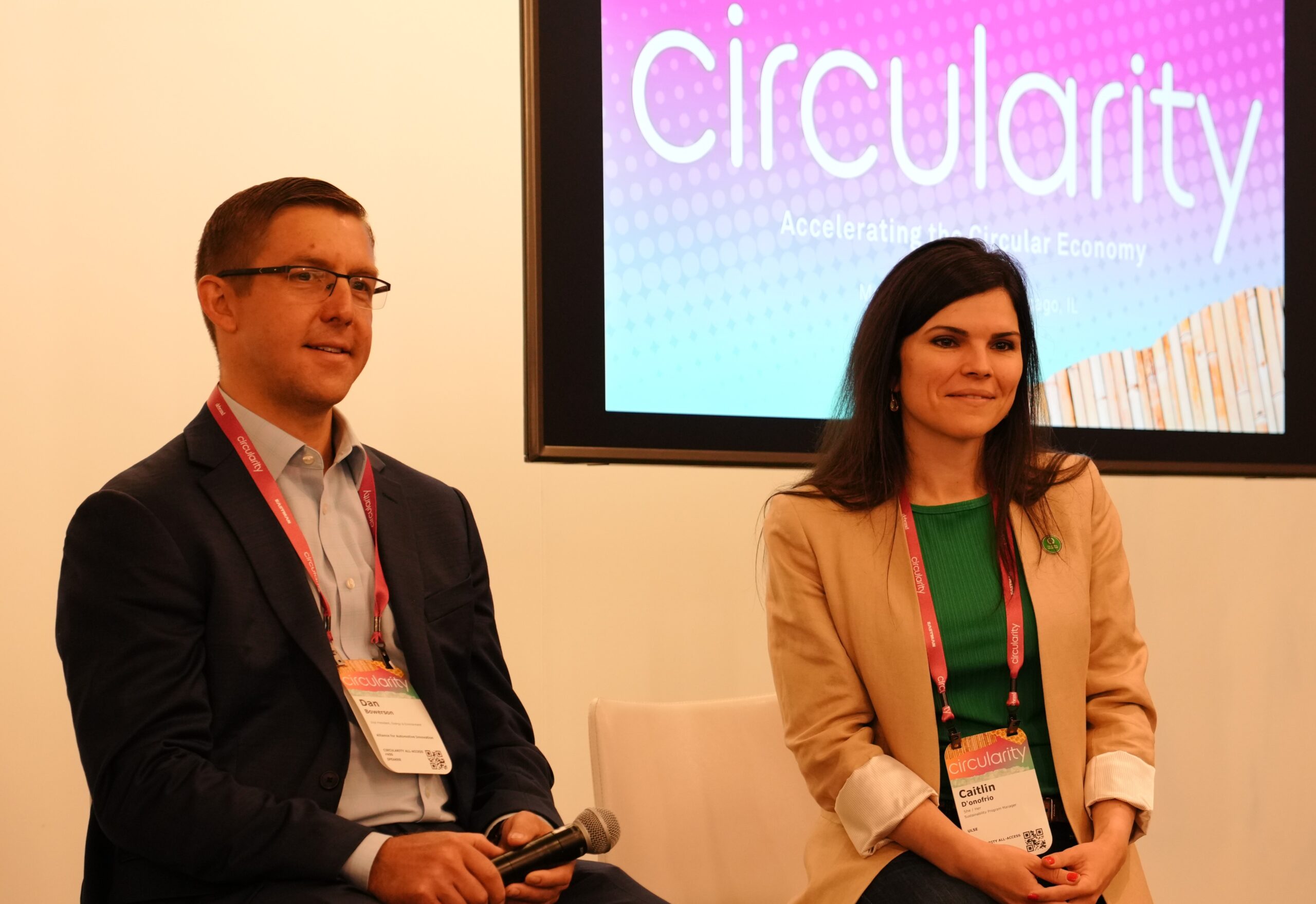 A man and a woman sit on a stage at a conference. Both wear name badges and lanyards. The woman smiles, and the man holds a microphone. Behind them, a screen displays the word "Circularity" in large letters.